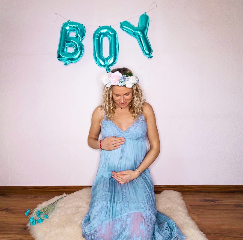 pregnant woman sitting in blue hippie dress underneath foil balloons spelling boy